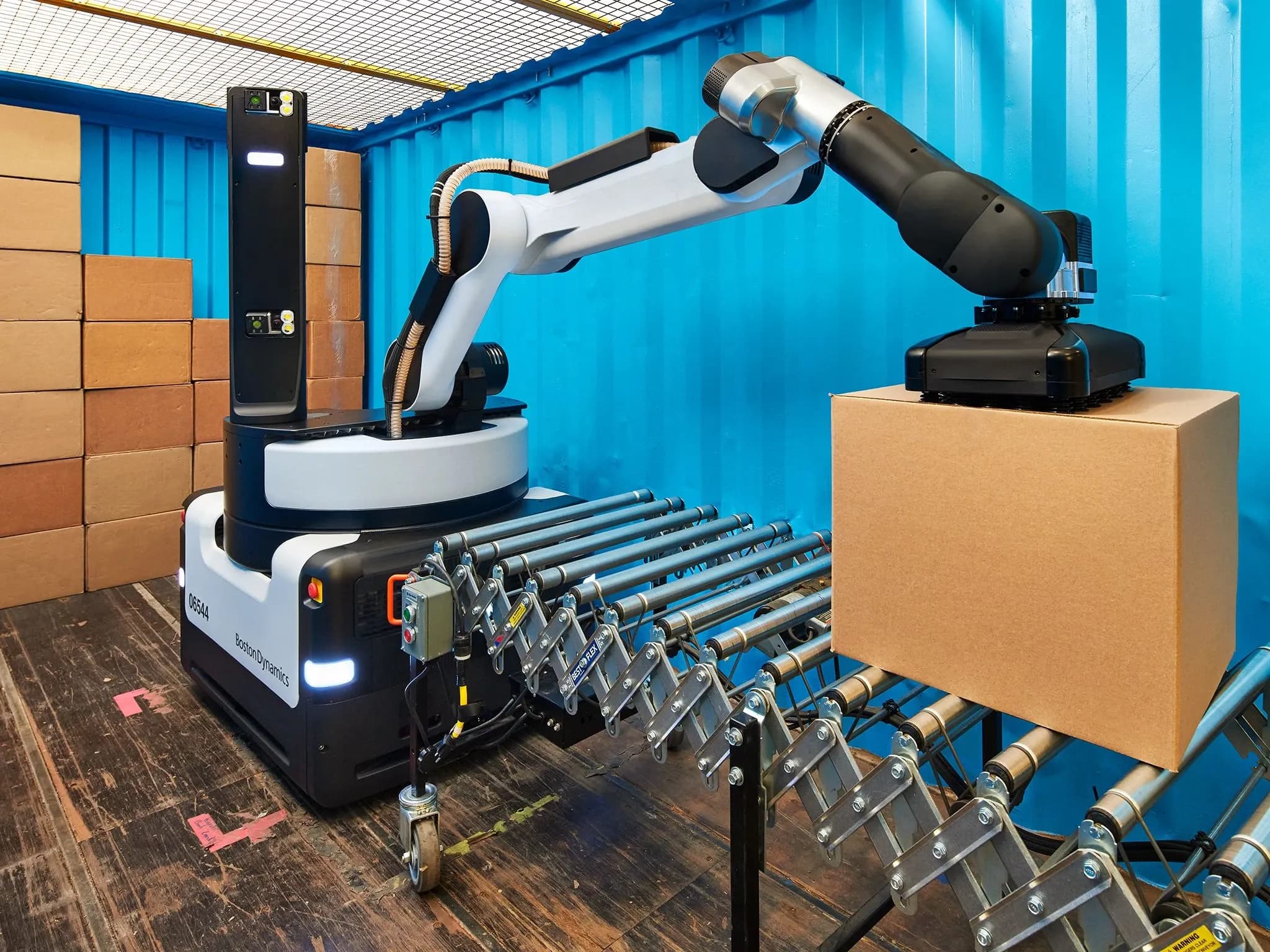 Робот Stretch от Boston Dynamics: крупная чёрно-серая мобильная база с шарнирным манипулятором и вакуумным захватом, снимающим картонную коробку с конвейера. На заднем плане — стопка коробок.