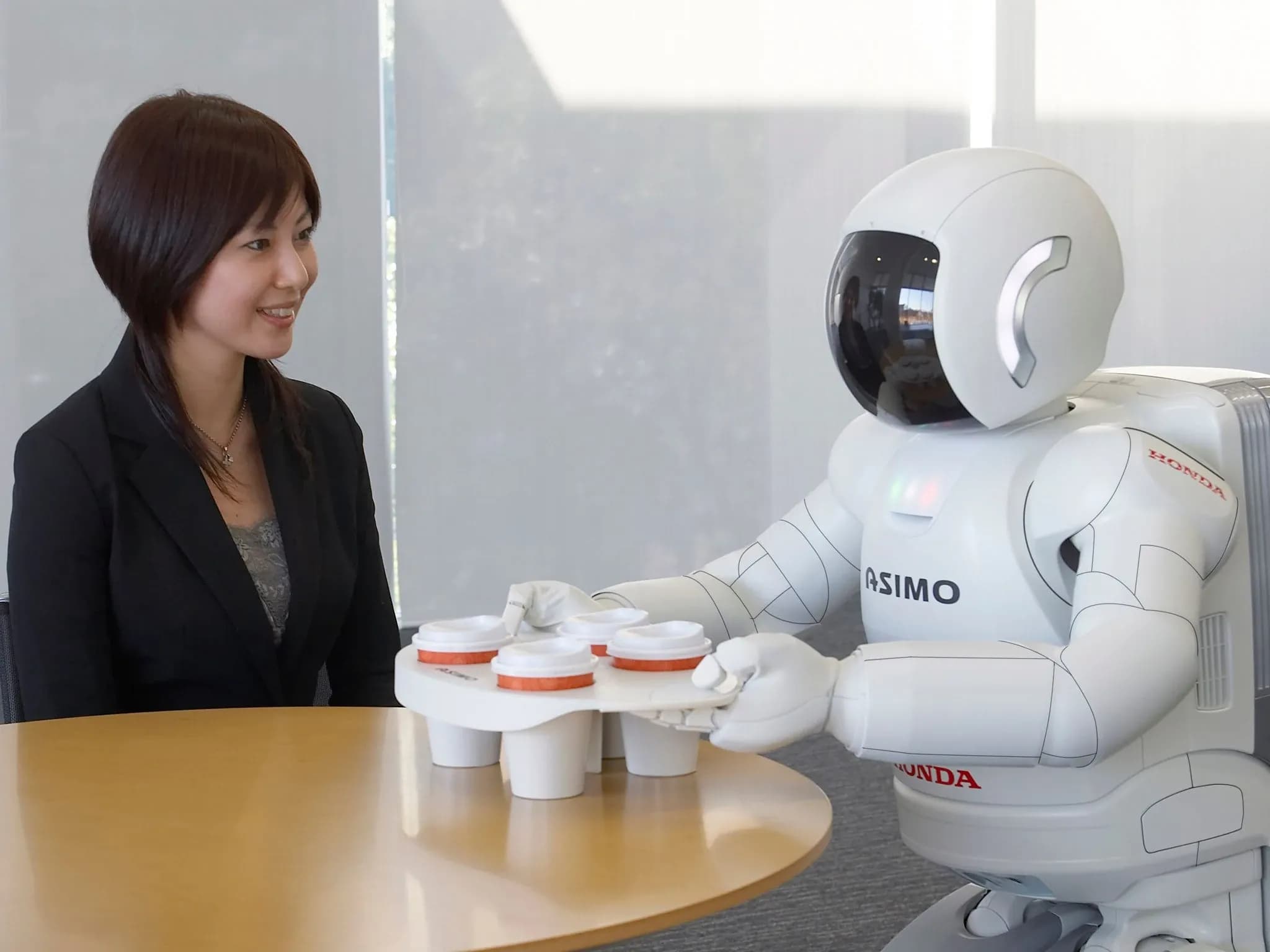 Женщина сидит за столом, пока робот Asimo подносит подставку с четырьмя чашками кофе.