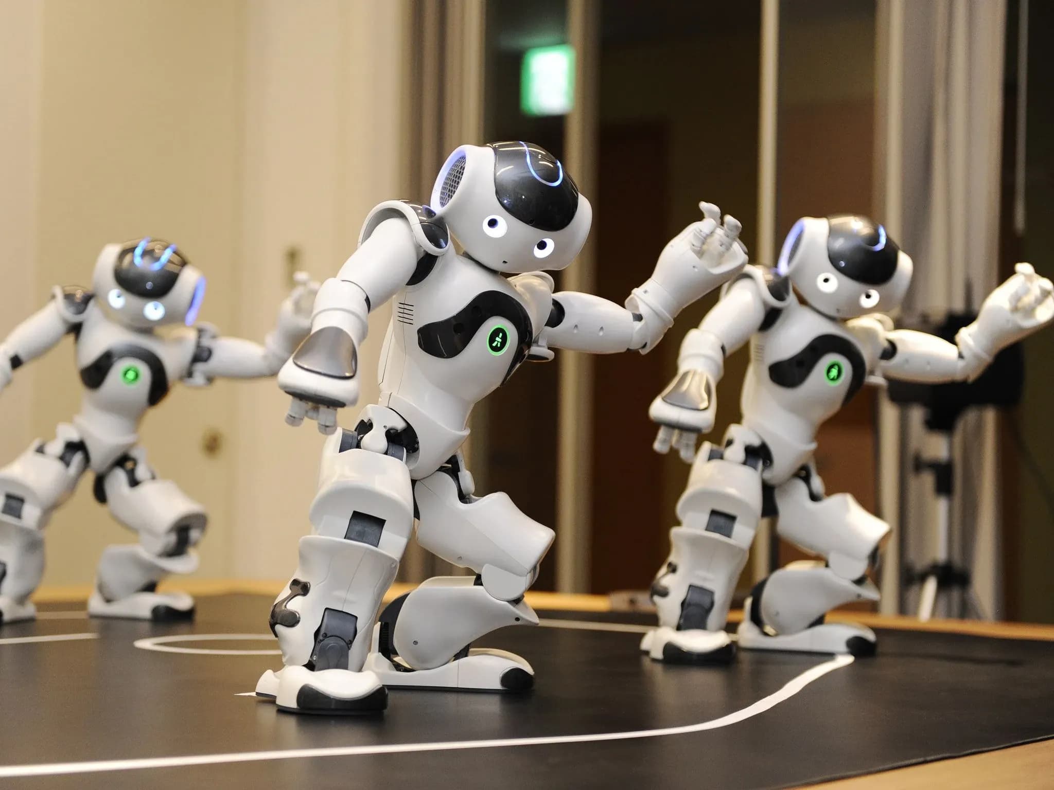 Три белых с серебристым гуманоидных робота Nao исполняют синхронные танцевальные движения.