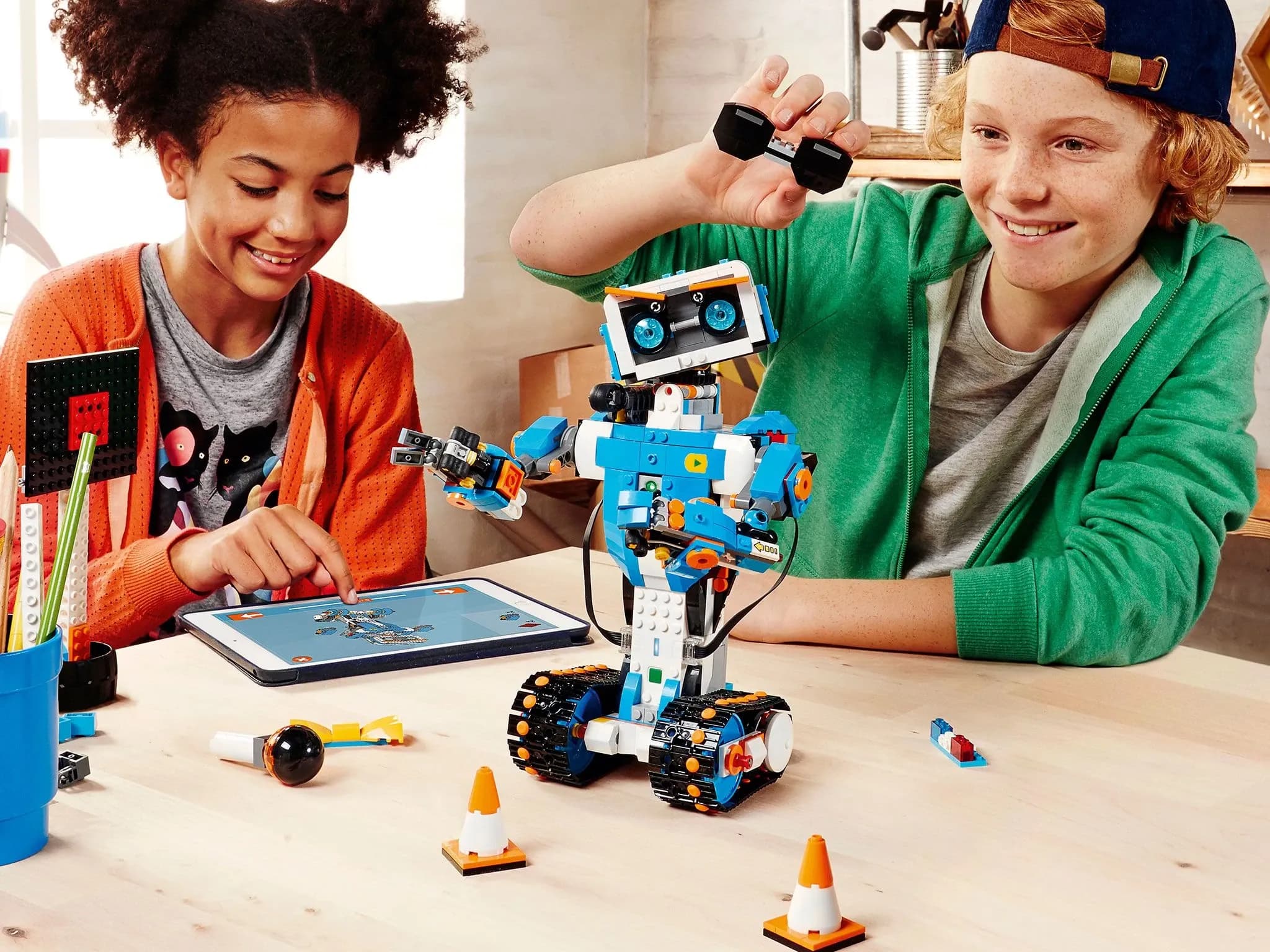 Двое подростков используют планшет для программирования робота из кубиков Lego: гуманоид с колёсными гусеницами вместо ног, двумя руками и милым лицом с голубыми глазами и оранжевыми бровями.