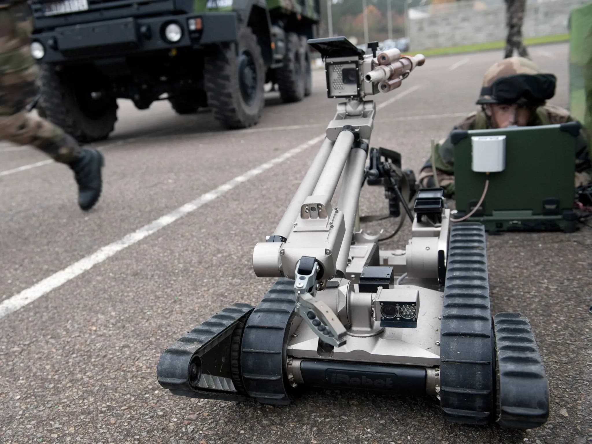 Военный лежит на улице перед ноутбуком. Перед ним — PackBot, сбоку — танк.
