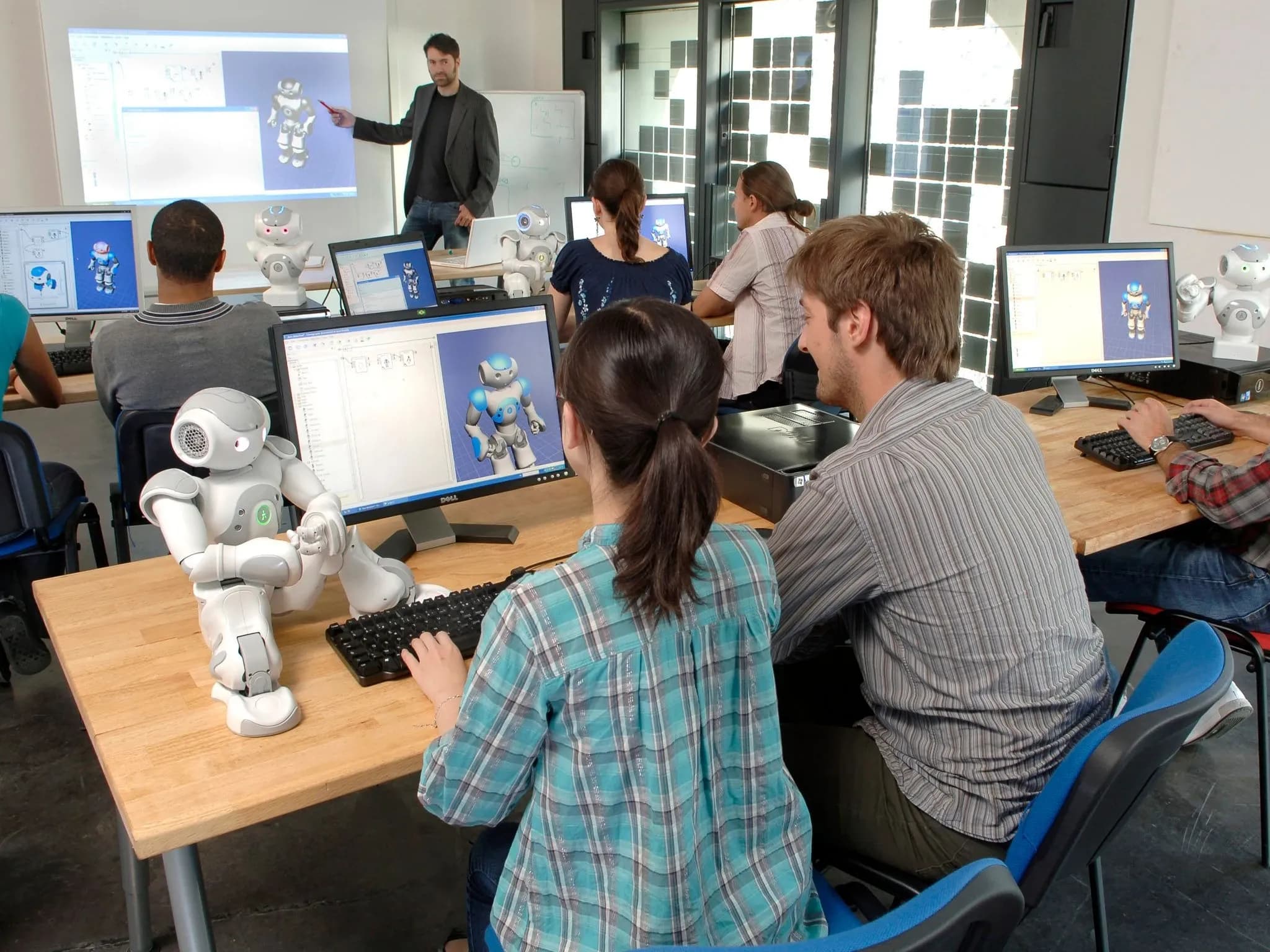 Компьютерный класс колледжа: студенты работают за столами с роботом Nao, на мониторе отображается их работа с роботом. Преподаватель у доски указывает на изображение Nao на экране.