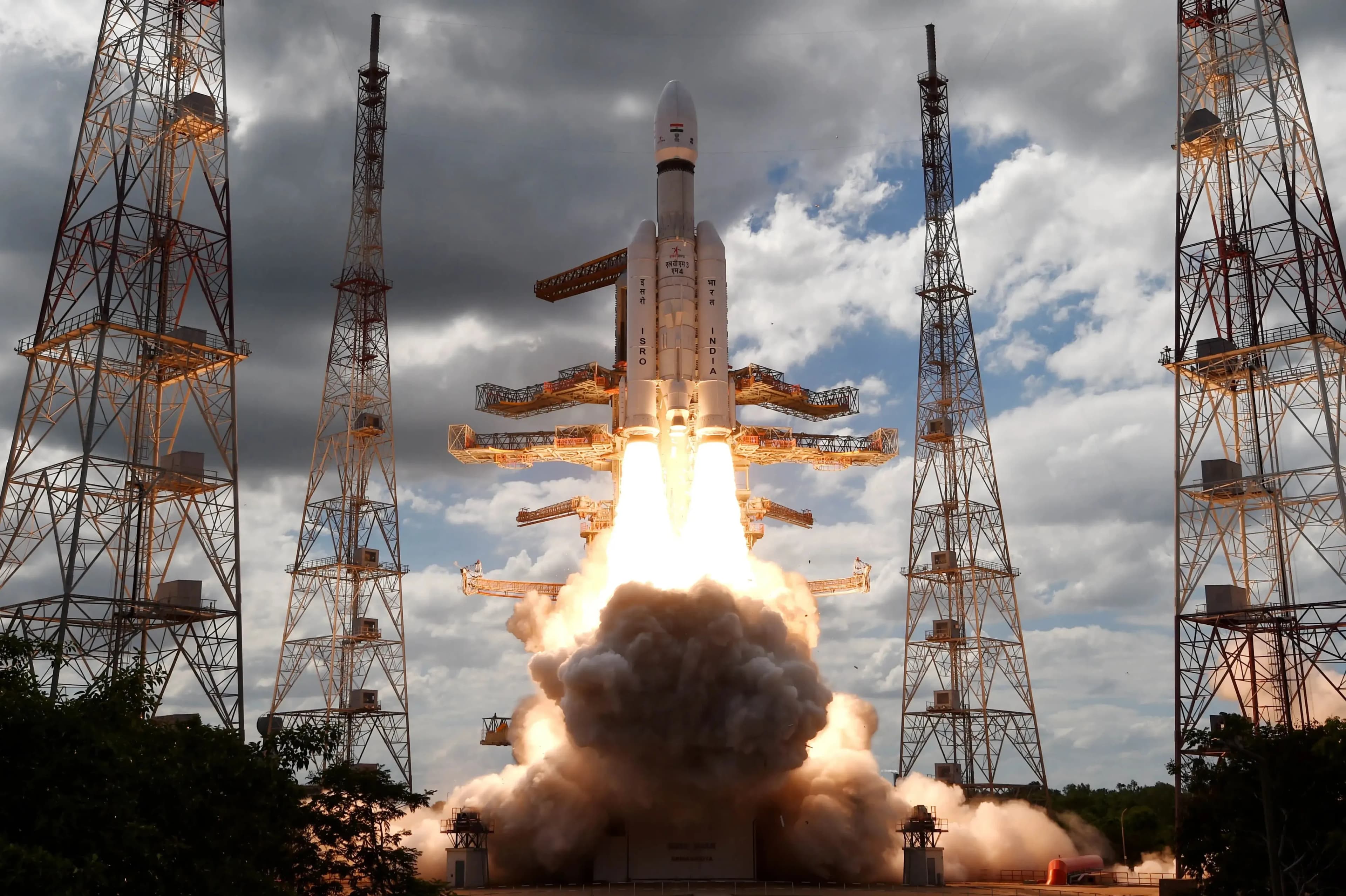 Запуск космического аппарата Chandrayaan-3: яркая жёлтая вспышка двигателей уносит его в частично облачное небо среди металлических антенных башен.