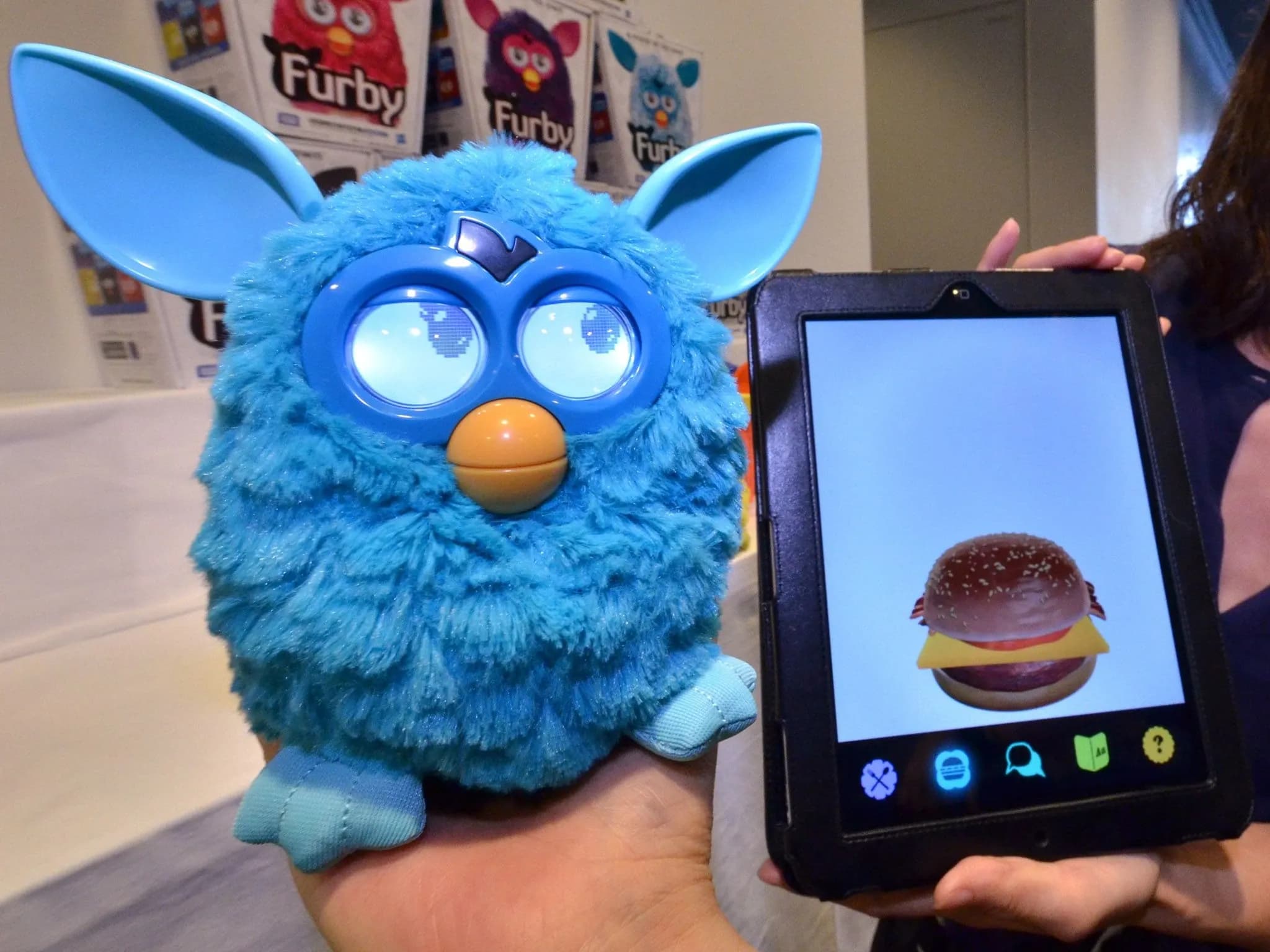 Синий пушисты Furby, похожий на сову, с большими глазами, жёлтым клювом и острыми ушами смотрит в камеру — рядом планшет с изображением чизбургера.