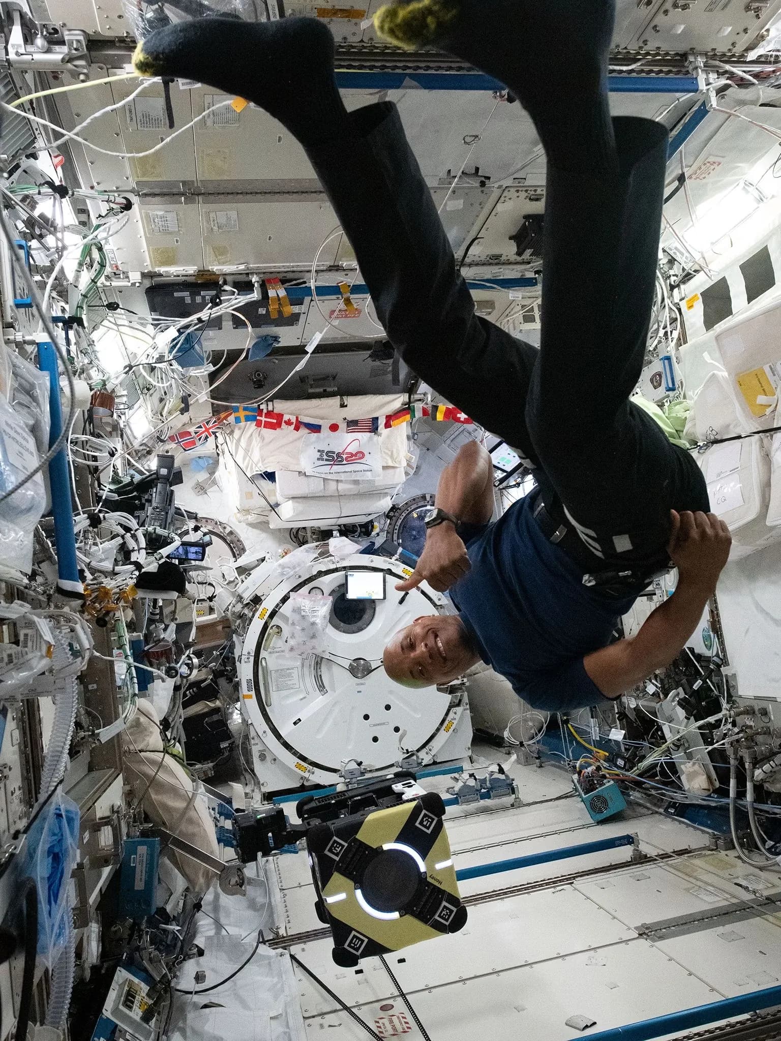 Улыбающийся астронавт вверх ногами парит рядом с роботом Astrobee на космической станции, окружённой электроникой.