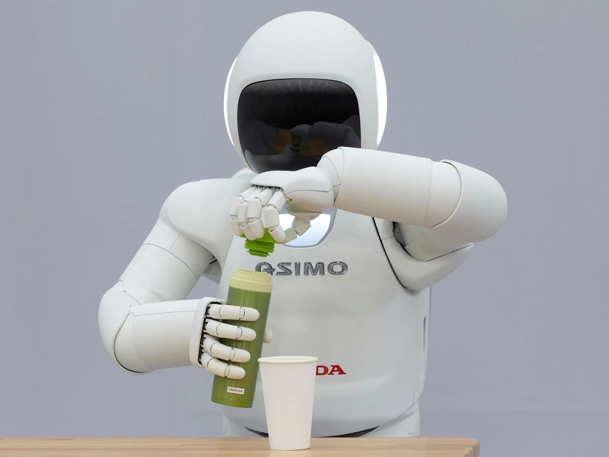 ASIMO от Honda открывает бутылку с зелёной жидкостью перед стаканом.