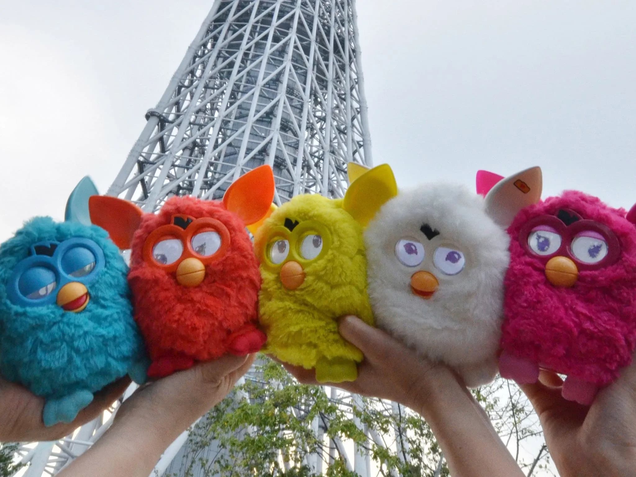 Две пары рук держат пять Furby — пушистых игрушек, похожих на сов, синего, красного, жёлтого, белого и розового цветов.