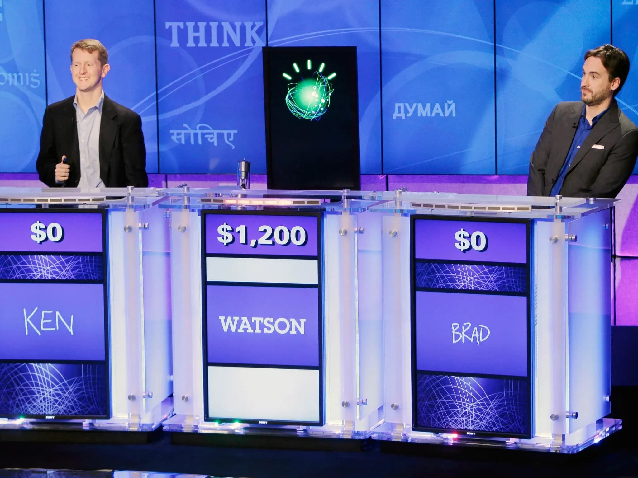 Двое участников и вертикальный экран с логотипом IBM Watson стоят за подиумами с табло, на которых отображаются суммы и имена игроков. У Watson — $1 200, у Кена и Брэда — по $0.